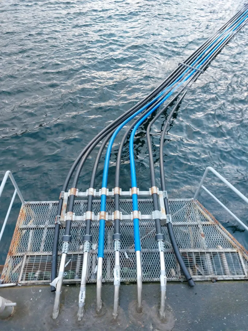 Vista desde arriba de las tuberías HDPE, sujetas con abrazaderas, bajando al agua junto a una pasarela metálica.