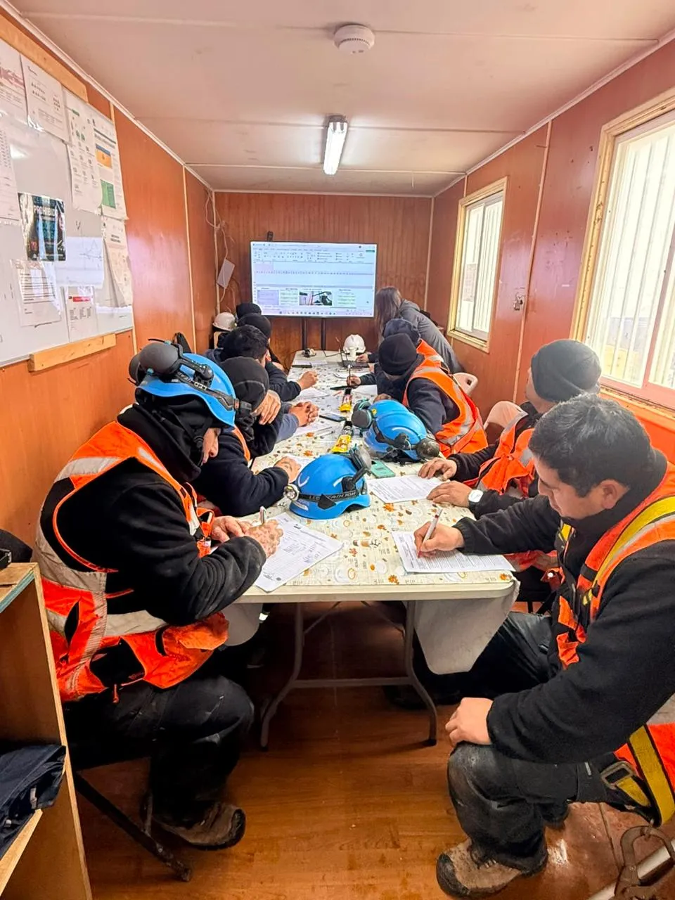 Técnicos con casco y chaleco naranja completando documentos en una mesa larga, dentro de la sala de una faena, con una pantalla al fondo.
