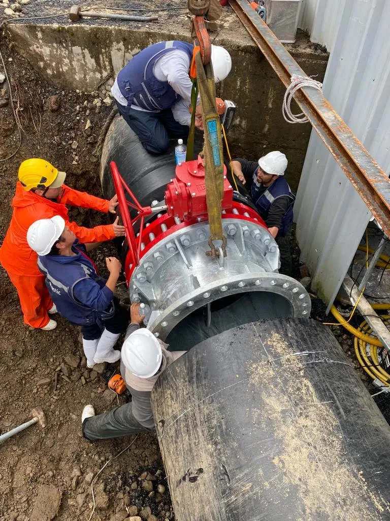 Cuadrilla con casco y buzo de trabajo bajando una válvula embridada roja sobre un tramo de tubería HDPE en una zanja.