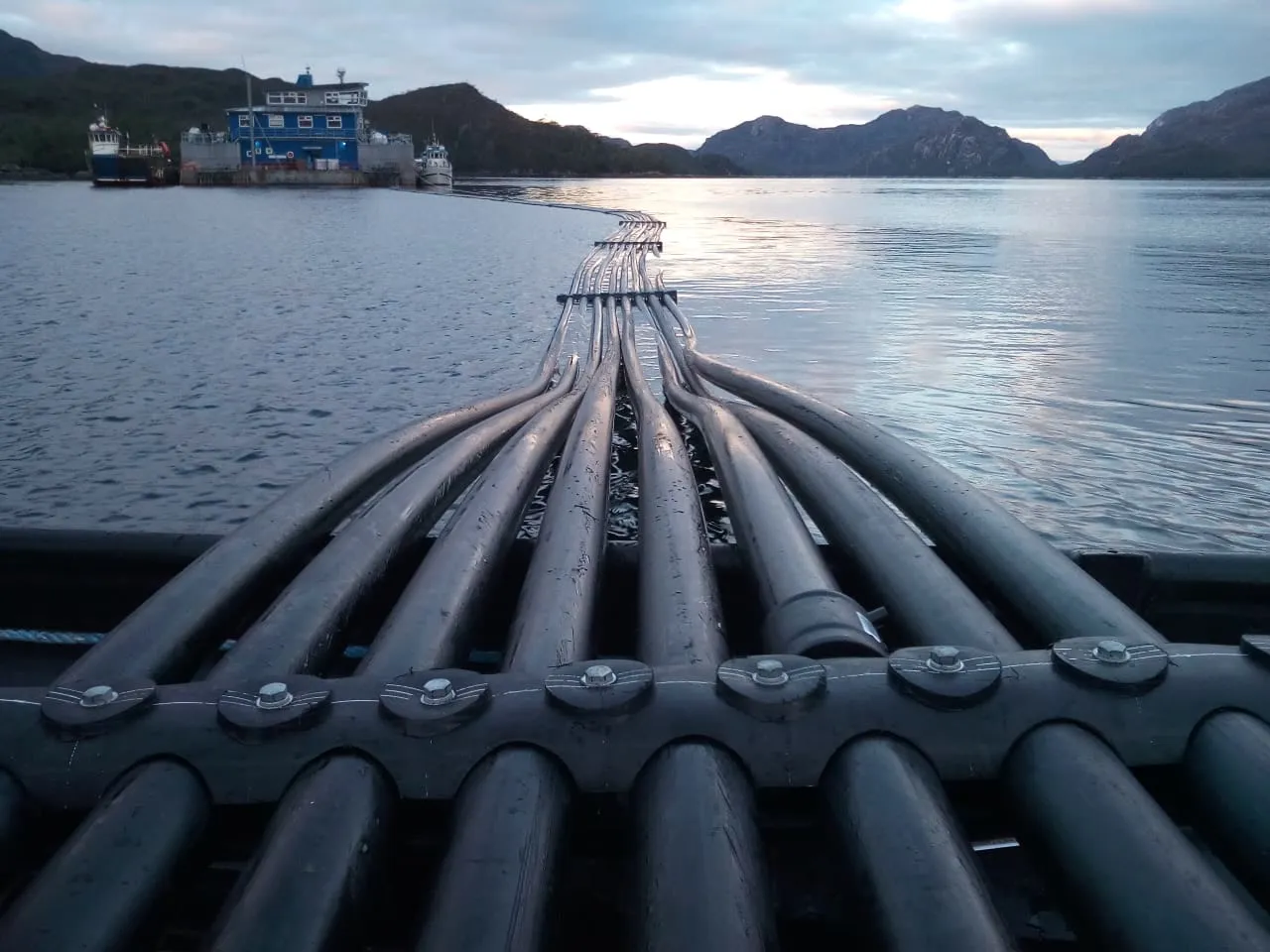 Haz de líneas de tubería HDPE tendidas sobre la pasarela de un pontón, avanzando por el agua hacia el fondo de un fiordo.