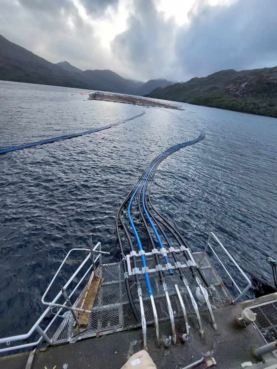 La línea de tubería HDPE saliendo hacia el fiordo desde la estructura de anclaje, con las montañas y el cielo nublado detrás.