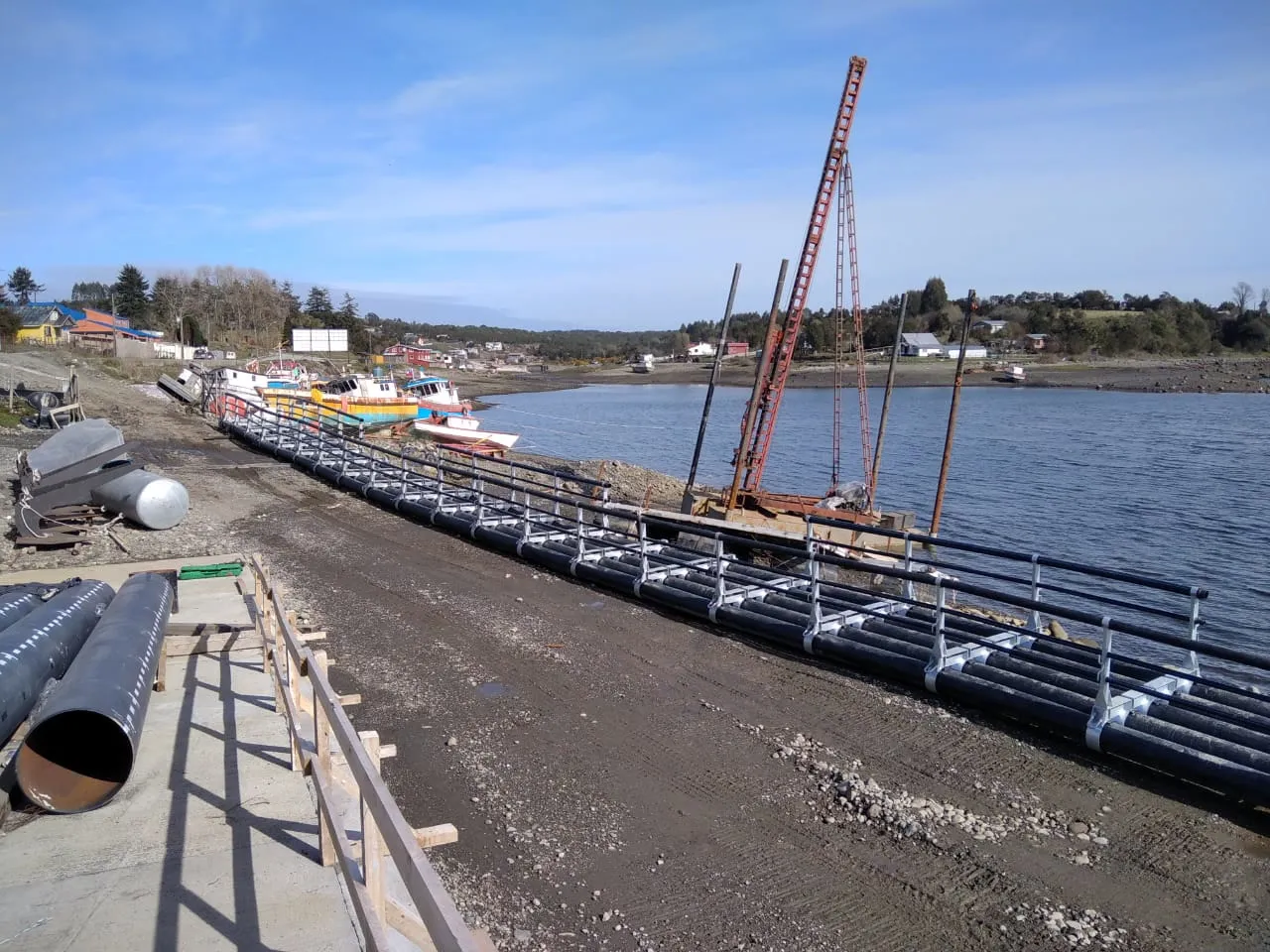 Línea de tubería HDPE montada sobre una pasarela de apoyo en la orilla de una caleta, con tubos acopiados en primer plano y el lago al fondo.